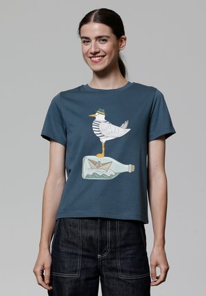 T-shirt en coton bleu présentant un graphisme d'une mouette en chemise rayée se tenant sur une bouteille en verre avec un bateau en papier à l'intérieur.