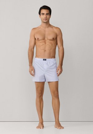 Boxers a rayas azules y blancas, tela ligera, cintura elástica y una etiqueta de logo en la parte delantera. Modelo de pie descalzo sobre un fondo liso.