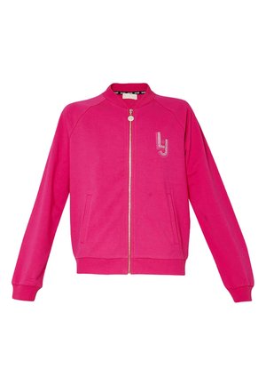 Personne portant une veste zippée rose vif avec un logo en strass sur le côté gauche de la poitrine et la main dans la poche latérale, sur un fond uni.