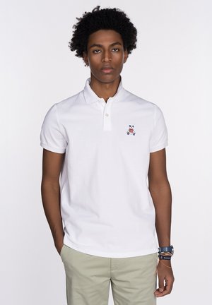 Camisa polo blanca de manga corta con una tapeta de tres botones y un pequeño logo bordado de un oso en el lado izquierdo del pecho. Textura de algodón suave.