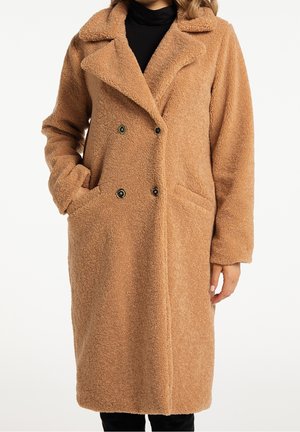 Femme portant un long manteau teddy marron clair à double boutonnage avec col large, poches latérales et vêtement intérieur noir sur fond blanc.