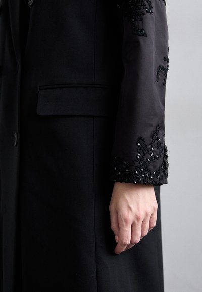 Melnā mētā ar pielāgotu dizainu, kurā ir izšūta piedurkne. Piedurkne ir dekorēta ar sekvīnēm un mežģīnēm, izceļot tekstūru un detaļas.