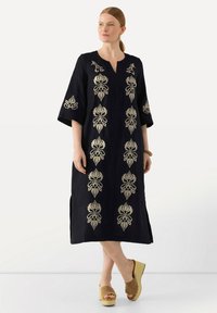 Schwarzes Leinenkleid mit goldener Blumenstickerei, V-Ausschnitt, Dreiviertelärmel, seitliche Schlitze, kombiniert mit braunen Wedgesandalen.