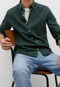 Man wearing dark green button-up shirt over white tee and light blue jeans sitting on wooden chair with black metal legs.