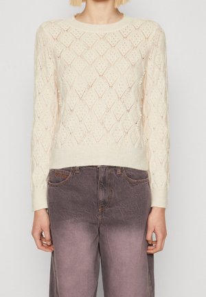 Femme portant un pull en maille crème à motif losanges avec des détails en perles et un jean gris délavé taille haute, debout devant un fond uni.