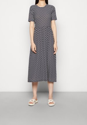 Femme portant une robe midi bleu marine avec un motif géométrique de losanges blancs et des sandales blanches tressées à enfiler, debout devant un fond uni.