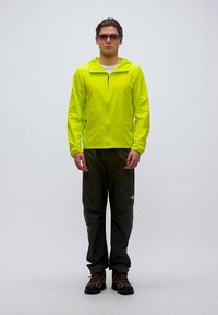 Άνδρας φοράει φωτεινό κίτρινο μπουφάν, μαύρο παντελόνι, παπούτσια πεζοπορίας και γυαλιά ηλίου, στέκεται μπροστά σε απλό φόντο.