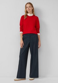 Czerwona swetrowa bluzka z długimi rękawami, zestawiona z ciemnymi, szerokimi spodniami w prążki. Prosty design z kontrastowym jasnym kołnierzykiem. Jasne buty.
