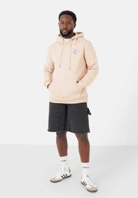 Felpa beige con cordini regolabili e grafica ricamata; abbinata a pantaloni shorts in denim neri e calze bianche, con dettagli a righe e sneakers.