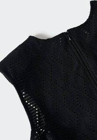 Vêtement noir sans manches avec des motifs en dentelle texturée en zigzag et à pois, montrant un gros plan du haut du dos et de la zone de la fermeture éclair.