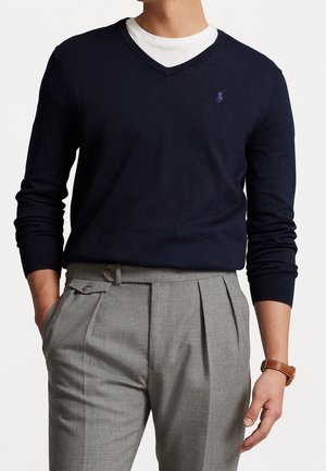 Mężczyzna w granatowym swetrze z dekoltem w serek na białej koszuli, szare plisowane spodnie i brązowy skórzany zegarek, ręce w kieszeniach.