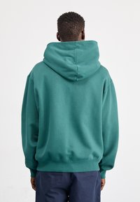 Osoba odwrócona plecami, ubrana w turkusową bluzę z kapturem i ciemnoniebieskie spodnie, na tle jednolitego białego tła.