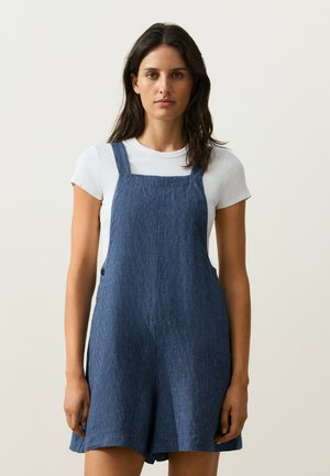 Frau mit langen dunklen Haaren, die ein weißes T-Shirt und einen blauen ärmellosen Cordoverall vor einem schlichten hellen Hintergrund trägt.