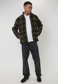 Młody mężczyzna z rękoma w kieszeniach, ubrany w zieloną koszulę z fleecu w kratę, białą koszulkę, ciemne dżinsy i czarne sneakersy, stojący na białym tle.