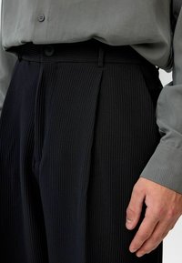 Zwarte ribbroek met plooien aan de voorkant, gedragen met een grijze lange mouw shirt. Details zijn onder andere een knoopsluiting en een brede tailleband.
