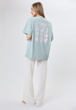 Kobieta stojąca tyłem, ubraną w jasnoniebieski oversize T-shirt z różowymi owocami i napisem "Farmers Market", w połączeniu z białymi szerokimi koronkowymi spodniami.