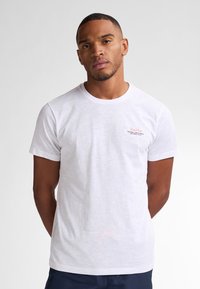 T-shirt bianco in cotone con maniche corte, colletto girocollo e piccolo logo rosso sulla parte sinistra del petto. Texture morbida, vestibilità rilassata.