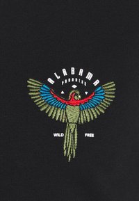 Tessuto nero con un design ricamato colorato di un pappagallo, che presenta dettagli verdi, blu e rossi. Il testo dice "PARADISO DELL'ALABAMA" e "LIBERO E SELVAGGIO."
