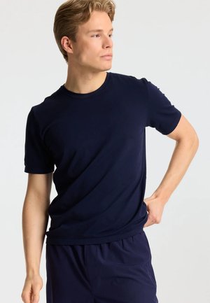 Lyshåret mand iført en tætsiddende navyblå t-shirt og bukser, kigger til siden med den ene hånd på hoften mod en ensfarvet baggrund.