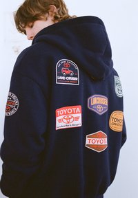 Tmavomodrá mikina s kapucňou s viacerými vyšitými odznakmi s logami Toyota a Land Cruiser v červenej, oranžovej a žltej farbe. Mäkká látka s voľným strihom.