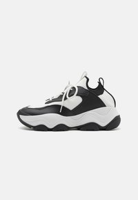 Zapatillas deportivas en blanco y negro con parte superior de malla texturizada, acentos de cuero, punta redondeada y suela de goma gruesa con surcos ondulados.
