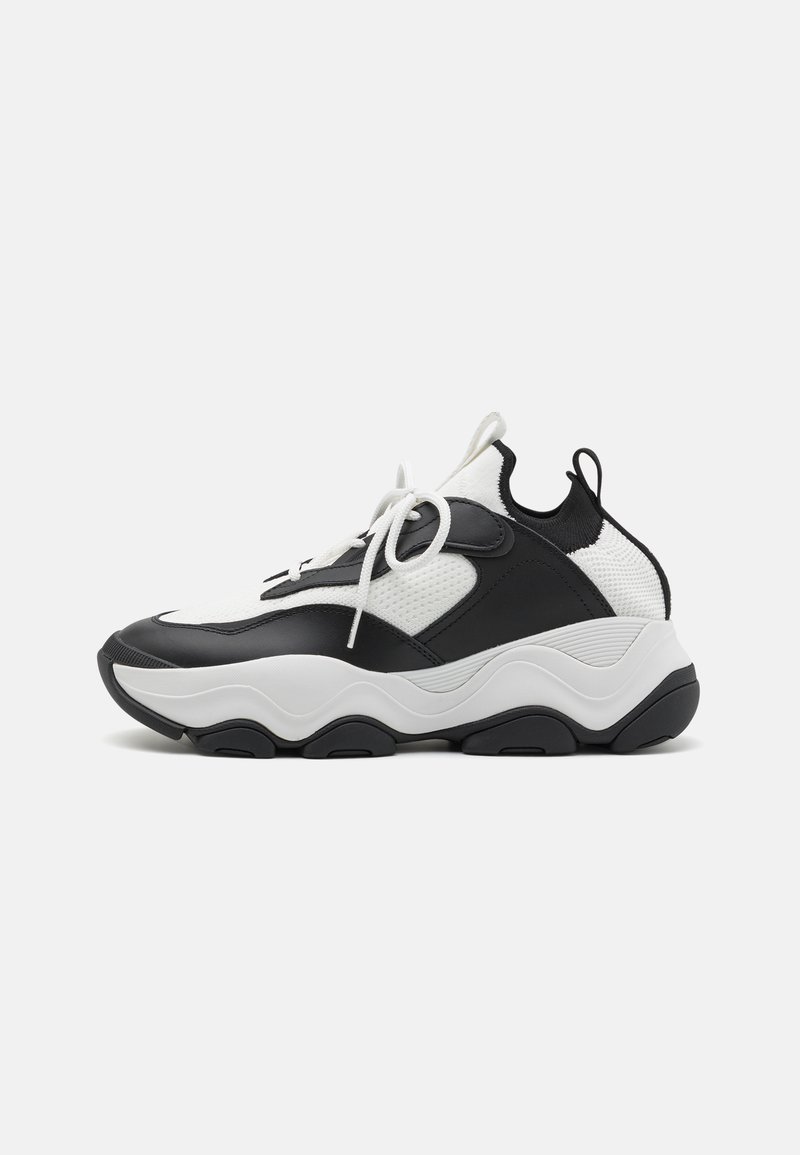 Zapatillas deportivas en blanco y negro con parte superior de malla texturizada, acentos de cuero, punta redondeada y suela de goma gruesa con surcos ondulados.