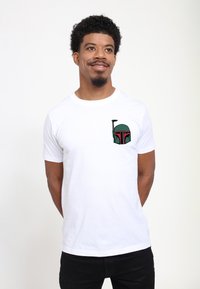 Hvid bomulds t-shirt med et grønt og rødt grafisk design af en hjelm på venstre bryst. Korte ærmer, rund halsudskæring og afslappet pasform.