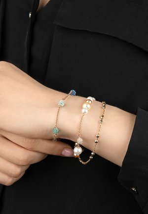 Trois bracelets autour d'un poignet : un avec des cristaux verts et bleus, un avec des perles blanches, et un autre avec des accents dorés et des perles, tous en design superposé.