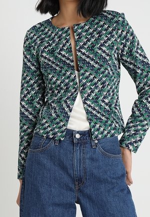 Kvinde iført en langærmet jakke med mønster i grøn, blå, hvid og sort over en hvid top og blå højtaljede jeans.