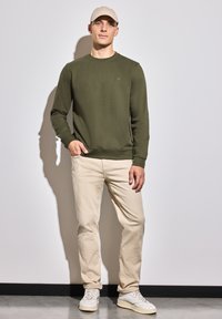 Zelená bavlněná mikina s kulatým výstřihem, zkombinovaná se světle béžovými kalhotami a bílými teniskami, na jednobarevném šedém pozadí.