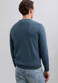 Hombre con cabello corto y rizado que lleva un suéter azul y jeans azul claro, de espaldas contra un fondo liso gris claro.
