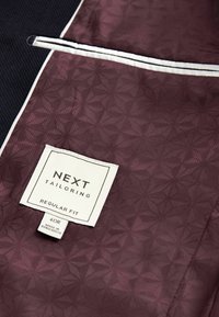 Bordeauxfarbene gemusterte Stoff mit einer geometrischen Textur, ausgestattet mit einem weißen Besatz und einem Etikett mit der Aufschrift "NEXT TAILORING, REGULAR FIT, 40R."