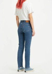 Jeans de mezclilla azul de cintura alta con un diseño ajustado, dos bolsillos traseros y una etiqueta de cuero marrón en la cintura; combinados con zapatillas de deporte azul claro.