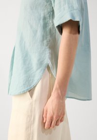 Personne portant une chemise bleu clair à manches courtes et un pantalon écru, vue de côté avec un focus sur le bras et les détails des vêtements.
