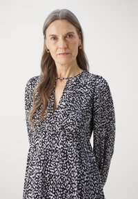 Valkoinen ja musta kuviollinen mekko, jossa on syvä V-pääntie, pitkät hihat ja tiukka vyötärö. Raskaasti tekstuurinen kangas ja laskeutuva hame.