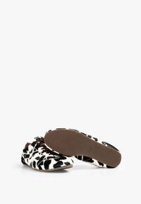 Zapatos tipo slip-on con estampado de vaca, parte superior de textil suave, cordones marrones y suela de goma texturizada. El diseño presenta un patrón en blanco y negro.