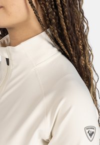 Krämfärgad jacka med hög krage och dragkedja. Har synlig sömnad och en logotypapplikation på vänster ärm. Slät textur.