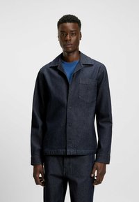 Mørk denimjakke med krave, brystlomme og frontknapper. Båret over en blå skjorte med lange ærmer og lige snit.