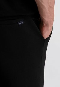 Zwarte joggers met een zijzak. Zacht stof met een gladde textuur. Bevat een label met de tekst "pierre cardin" op de tailleband.