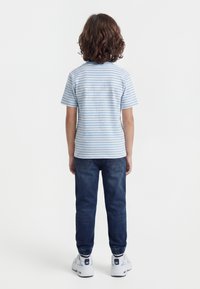 Keskmise pikkusega pruunide juustega laps, kes kannab sinise ja valge triibulise T-särgi, tumedate siniste teksade ja valgete tennistega, vaatega selja poole.