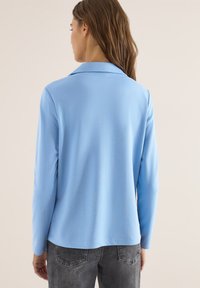 Femme aux longs cheveux bruns portant une chemise à manches longues bleu clair et un jean gris, vue de dos sur un fond uni.