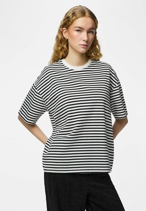 Jeune femme aux cheveux blonds ondulés portant un t-shirt à manches courtes rayé noir et blanc et un pantalon noir, les mains derrière le dos.