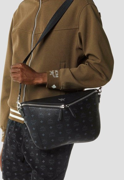 Man draagt een bruine jas met rits en zwarte broek met patroon, houdt een zwarte leren schoudertas met zilveren ritsen vast.