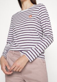 Långärmad t-shirt i ljusgrått med mörklila ränder. Har en liten orange logotyp på bröstet, rund halsringning och mjukt tyg.