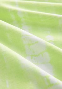 Tissu vert clair avec des motifs tie-dye blancs, présenté en plis doux mettant en valeur la texture et les variations de couleur.