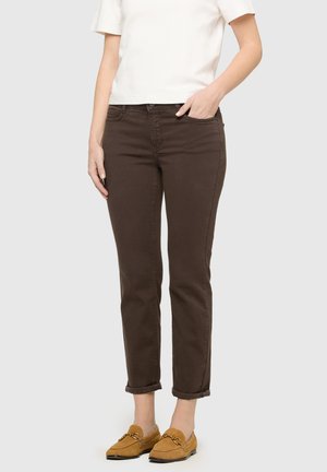 Vrouw die donkerbruine slim-fit broek, wit topje met korte mouwen en lichtbruine loafers draagt, staand met één hand in de zak.