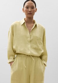 Camicia di lino giallo chiaro con colletto, bottone frontale e taschino sul petto; abbinata a pantaloni con vita regolabile dello stesso colore.