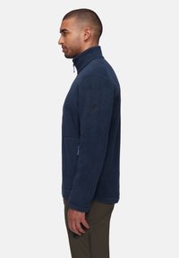 Navy fleece jacket with high collar, half-zip, zippered pocket, and logo patch on the sleeve. Soft texture and relaxed fit.