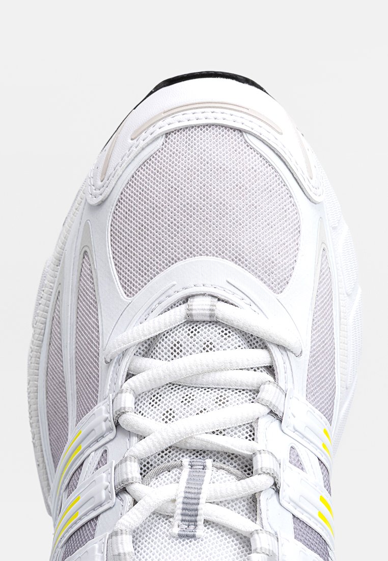 Zapatilla deportiva blanca con parte superior de malla, acentos grises y rayas amarillas brillantes. Cuenta con cordones robustos y una suela texturizada para mayor agarre.