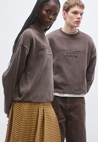 Sudadera marrón con el logo bordado "TOMMY JEANS", combinada con una falda amarilla a cuadros y plisada. Ambas prendas tienen diseños cómodos y relajados.